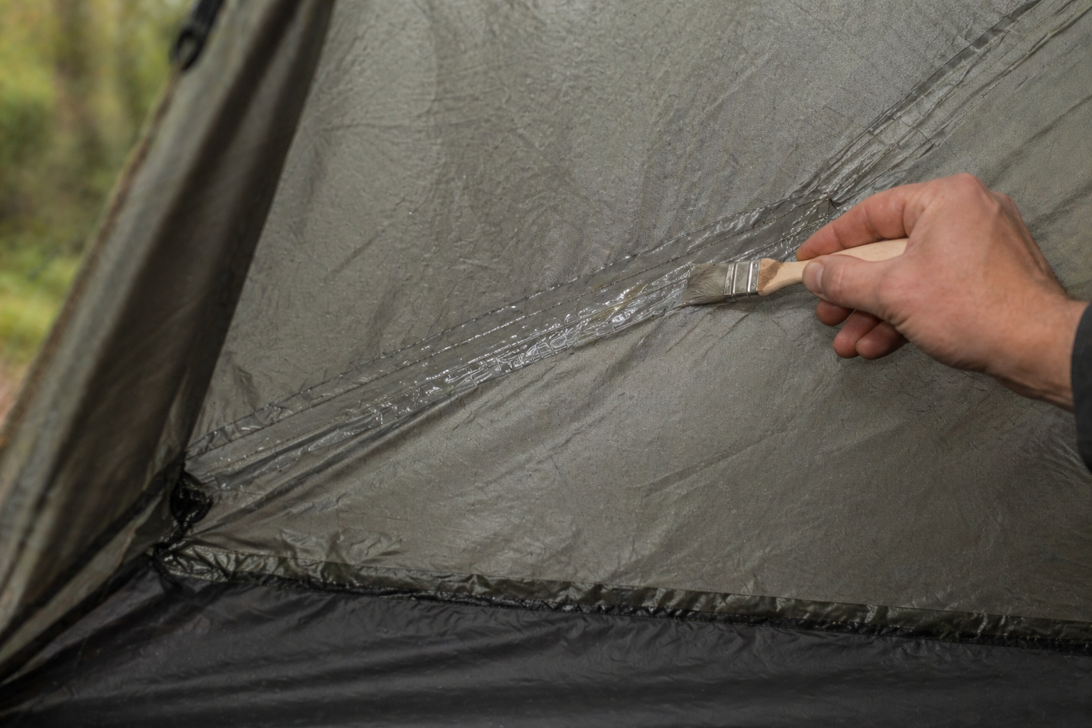 Close-up of a small paintbrush applying seam sealer along the interior side of a tent seam, with a visible glossy line of fresh sealer covering the stitching
